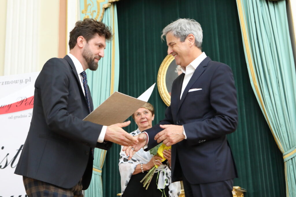 Zdjęcie. Mężczyzna o ciemnych włosach i brodzie podaję rękę do gratulacji siwemu, szczupłemu uśmiechniętemu mężczyźnie. W drugiej ręce trzyma otwartą teczkę.