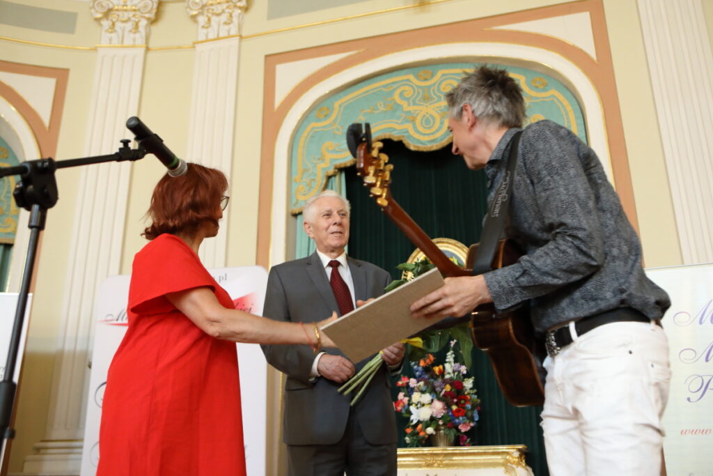 Zdjęcie. Ruda kobieta w czerwonej sukience podaje dłoń do gratulacji szczupłemu mężczyźnie z gitarą. Tn patrzy w kierunku starszego mężczyzny trzymającego bukiet słoneczników.