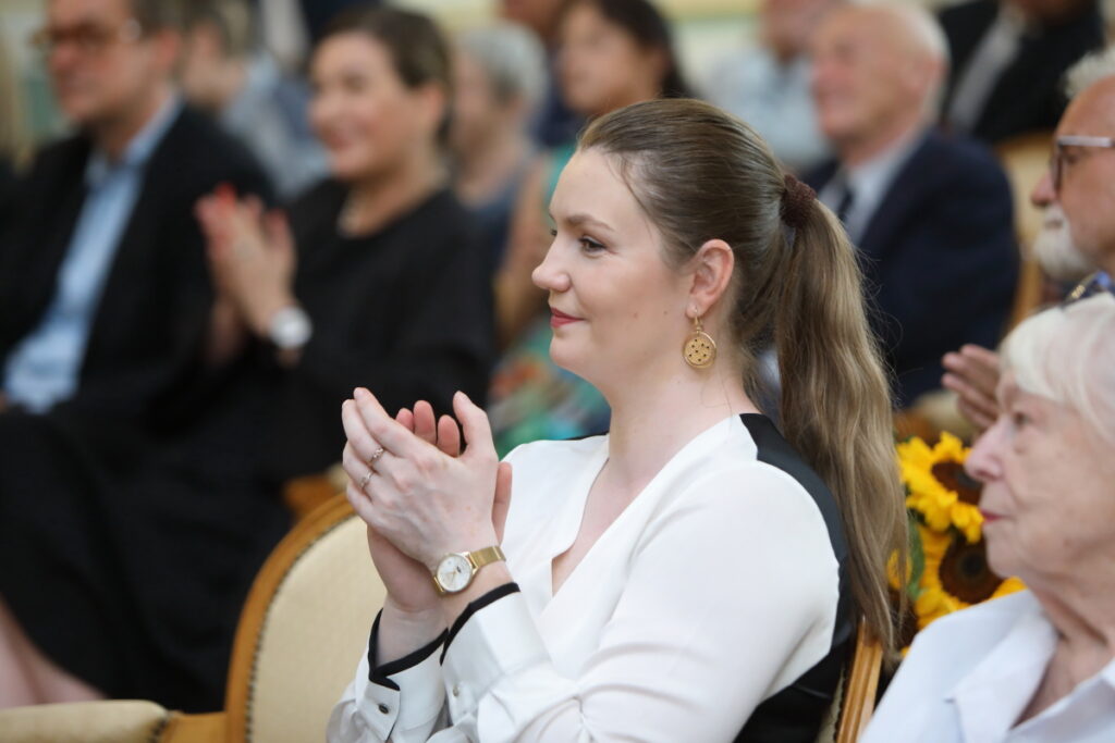 Zdjęcie. Blondynka o długich włosach, spiętych w kąski ogon ubrana w bluzkę białą z przodu, a czarną na plecach siedzi na krześle i się uśmiecha. Ręce ma złożone jakby do oklasków. To dyrektor KCK Agata Klimczak-Kołakowska.