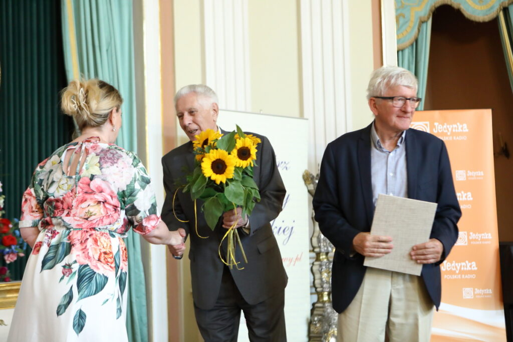 Zdjęcie. Kobieta w kwiecistej sukni odbiera gratulacje z rąk starszego mężczyzny. Ten w drugiej ręcce trzyma bukiet sloneczników. Obok stoi drugi starszy pan z płócienną twardą teczką.