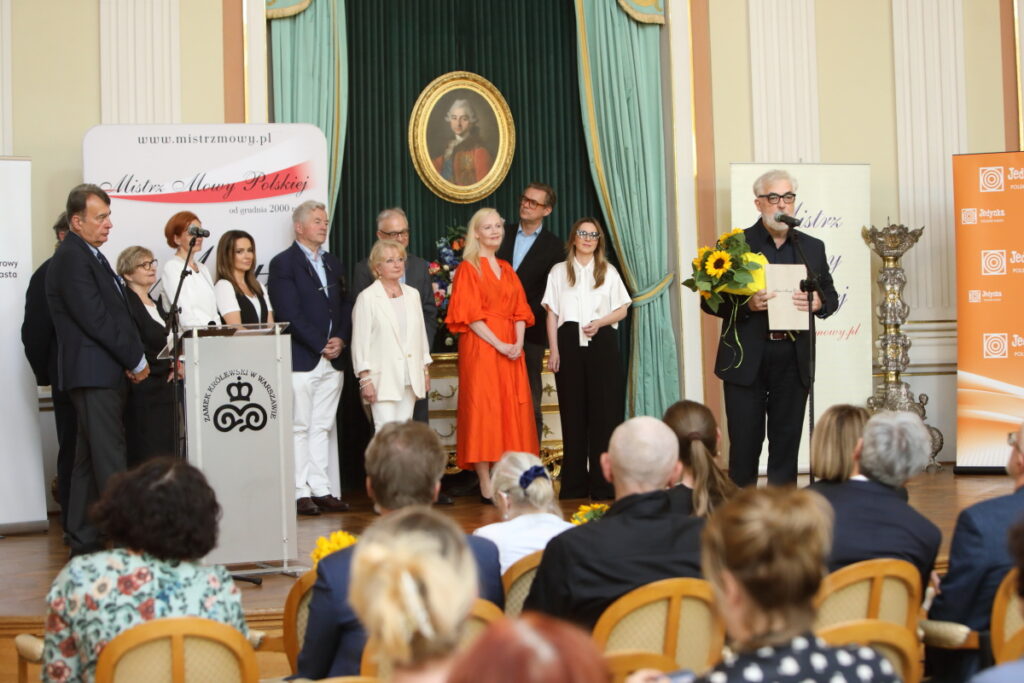 Zdjęcie. Grupa ludzi stoi w dwóch rzędach na scenie. W tle ciemnozielona zasłona, a na niej owalny portret Stanisława Augusta Poniatowskiego. Obok wysoki siw pan mówi do mikrofonu. Przzygląda się im widownia siedząca na historycznych krzesłach.