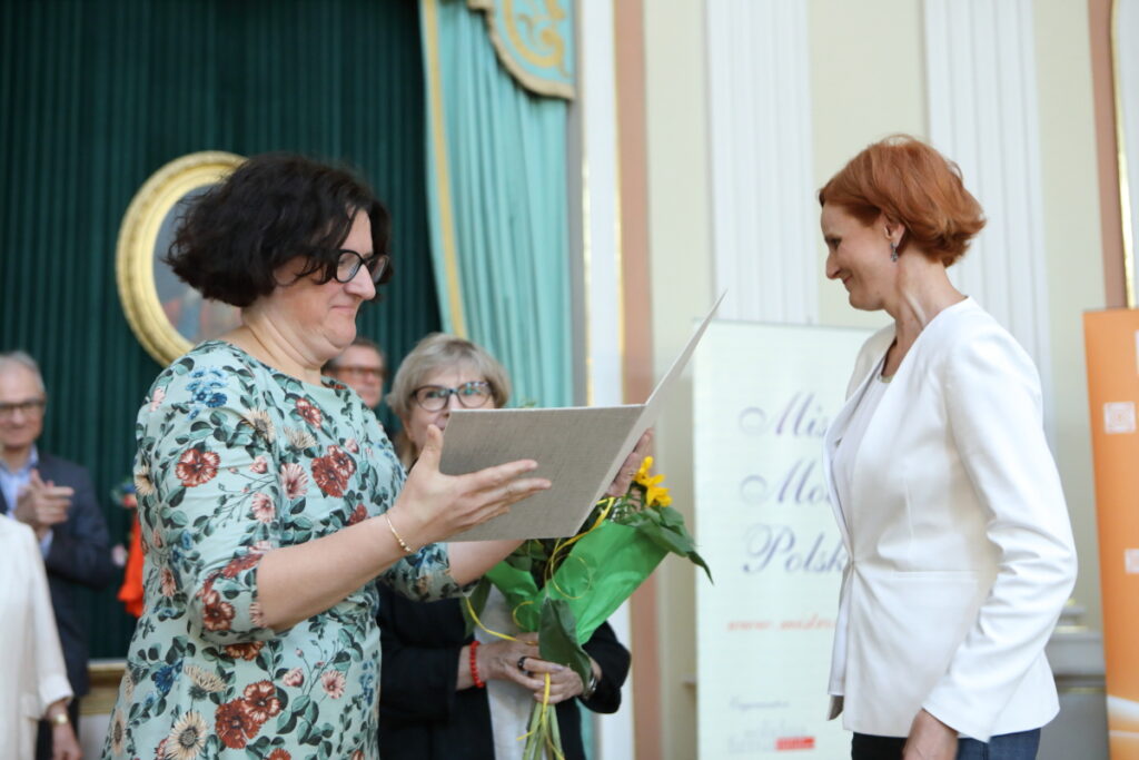 Zdjęcie. Kobieta w kwiecistej sukience o ciemnych włosach do ucha wręcza szczupłej rudej kobiecie w białej marynarce otwartą jasną teczkę.