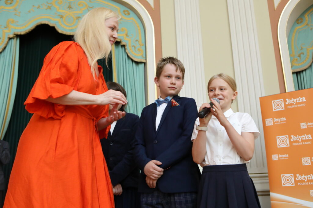 Zdjęce. Kobieta w pomarańczowej sukience o włosach blond patrzy w kierunku mówiącej do mikrofonu dziewczynki o włosach blond. Obok dziewczynki w rzędzie stoi dwójka dzieci.