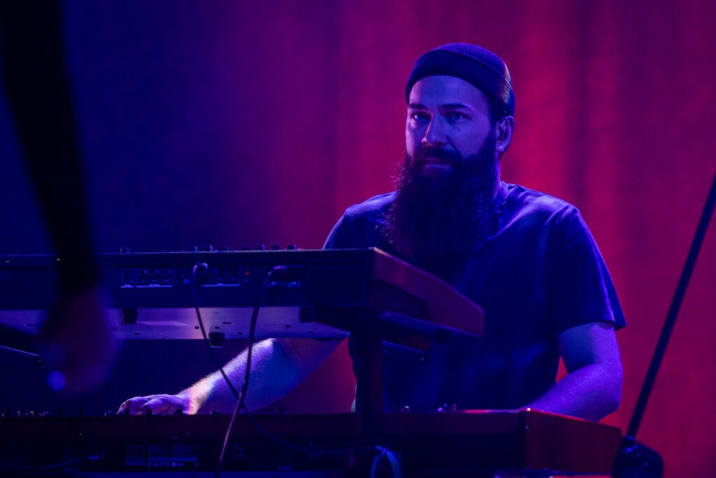 Zdjęcie. Mężczyzna z długą ciemną brodą i dopasowanej czapce na głowie gra na keyboardach. W tle bordowa kotara. Mężczyzna jest oświetlony niebieskim reflektorem.