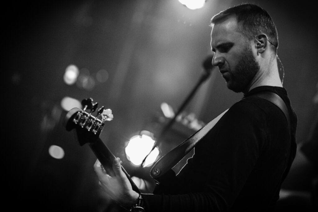 Czarno-białe zdjęcie. Krótko obcięty mężczyzna z brodą gra na gitarze.