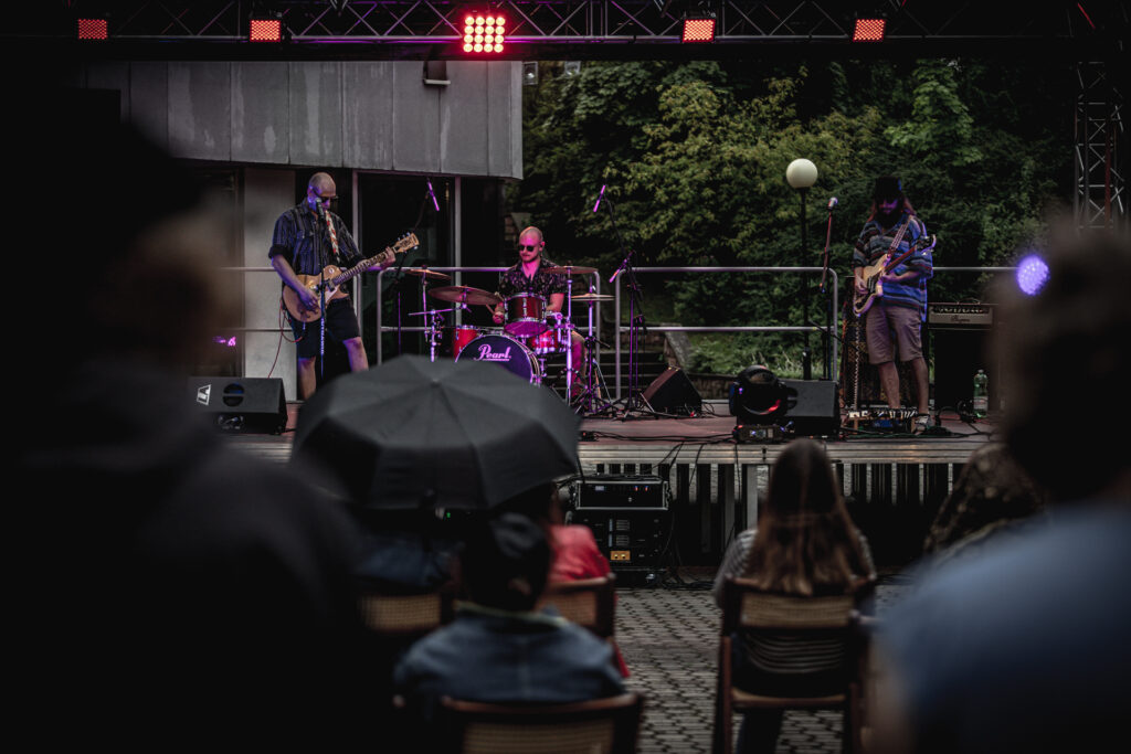 Zdjęcie. Koncert na Letniej Scenie KCK. Na scenie trzech muzyków: dwóch gitarzystów i perkusista.