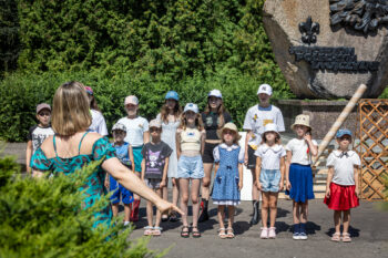 Zdjęcie. Grupa dzieci stoi w dwuszeregu. Przed nimi instruktorka. Z nimi pomnik Harcerzom poległym za ojczyznę.