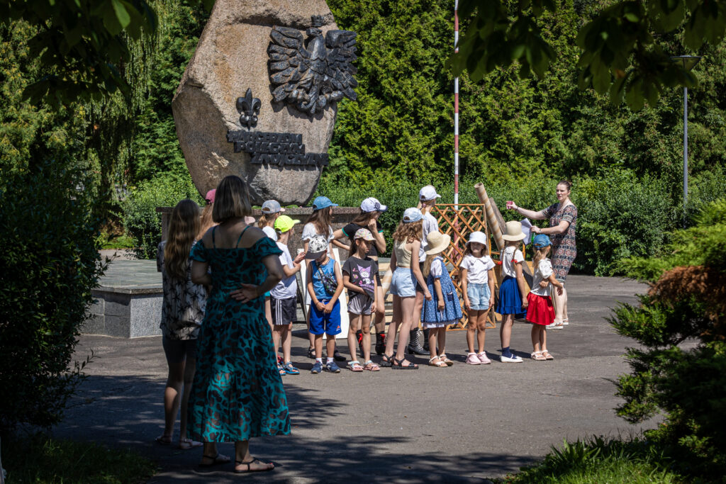 Zdjęcie. Dzieci stoją w dwóch rzędach oglądają się w kierunku instruktorki stojącej w pobliżu konstrukcji.