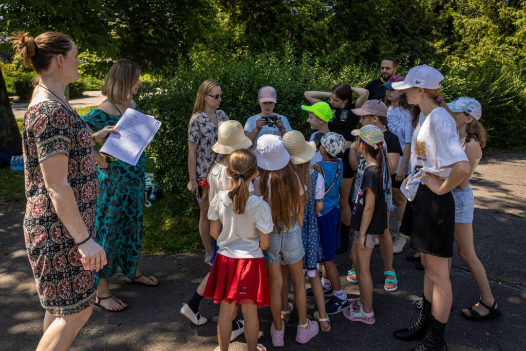 Zdjęcie. Grupa dzieci stoi w kółku blisko siebie. Patrzą w kierunku chłopca, który w rękach trzyma stary aparat fotograficzny. Obok stoją trzy instruktorki.