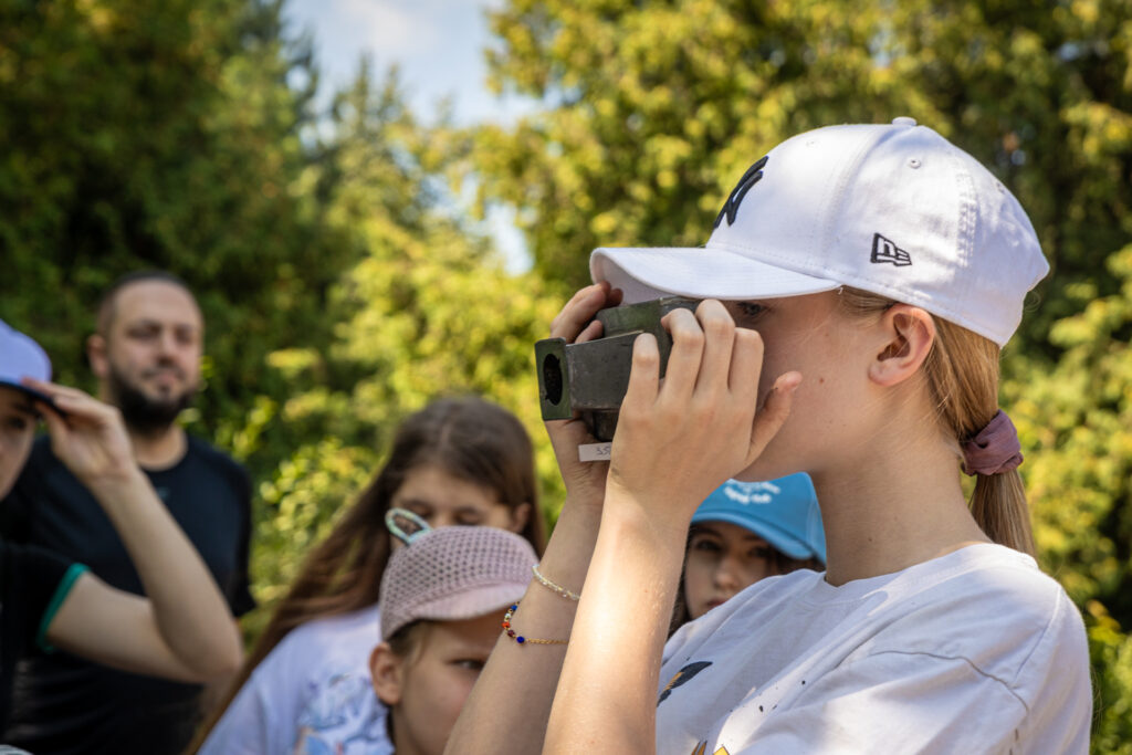 Zdjęcie. Dziewczynka w białej czapce bejsbolówce przykłada do oka stary metalowy aparat.