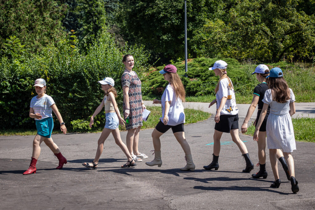 Zdjęcie. Dzieci maszerują jedno za drugim po okręgu. Obok stoi instruktorka.