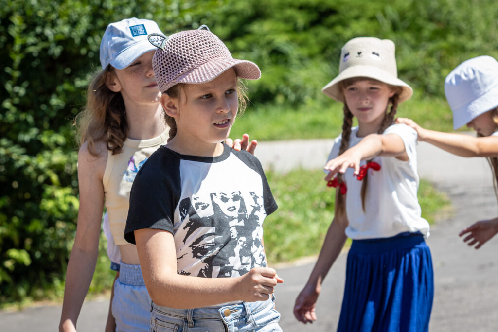 Zdjęcie. Grupa dziewczynek chodzi w kole jedna za drugą. Każda trzyma swoją poprzedniczkę lewą ręką za lewe ramię.