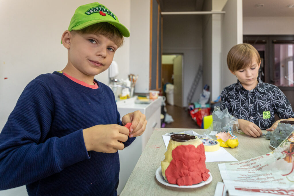 Zdjęcie. Chłopiec w groszkowej bejsbolówce patrzy w stronę aparatu fotograficznego. W dłoniach trzyma czerwoną plastelinę. Przed nim wulkan oklejony czerwoną i żółtą plasteliną.