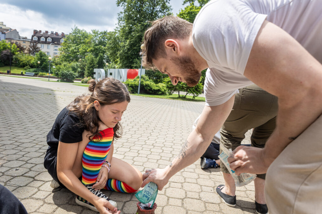 Zdjęcie. Mężczyzna nasypuje do kubka oblepionego plasteliną sodę oczyszczoną z niewielkiego opakowania. Przygląda się temu nastolatka w kolorowych, tęczowych getrach.