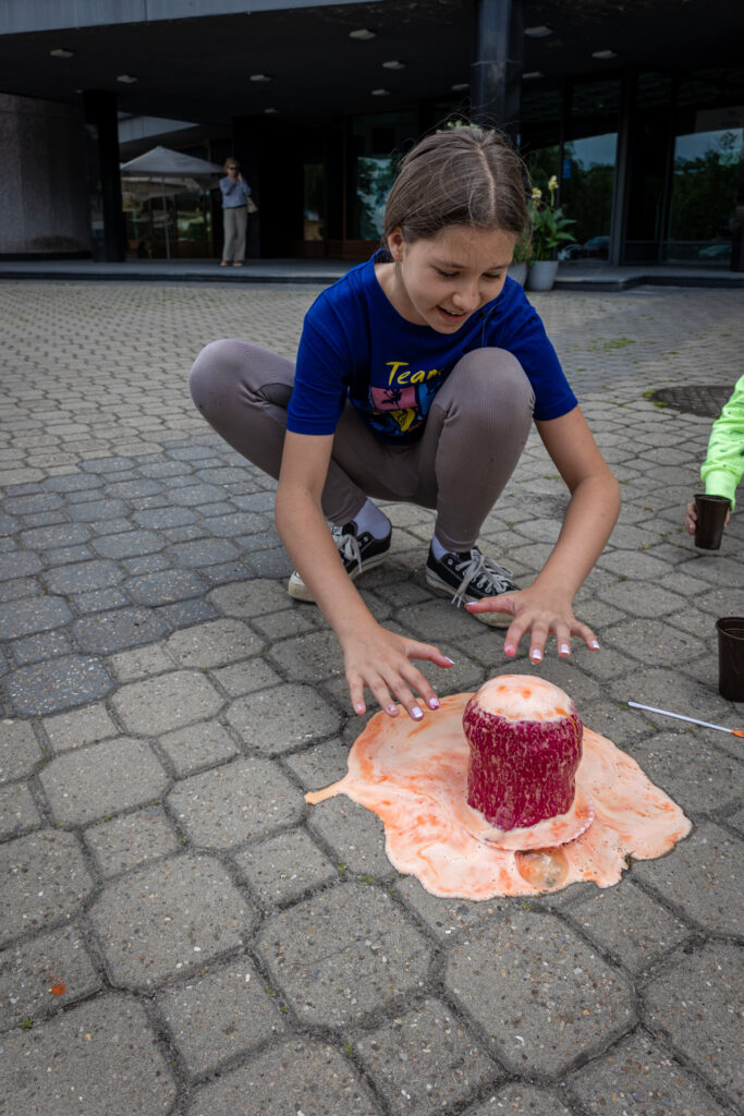 Zdjęcie. Ciemnowłosa nastolatka z radością i uwagą obserwuje pomarańczową pianę, która wylewa się z jej bordowego wulkanu.