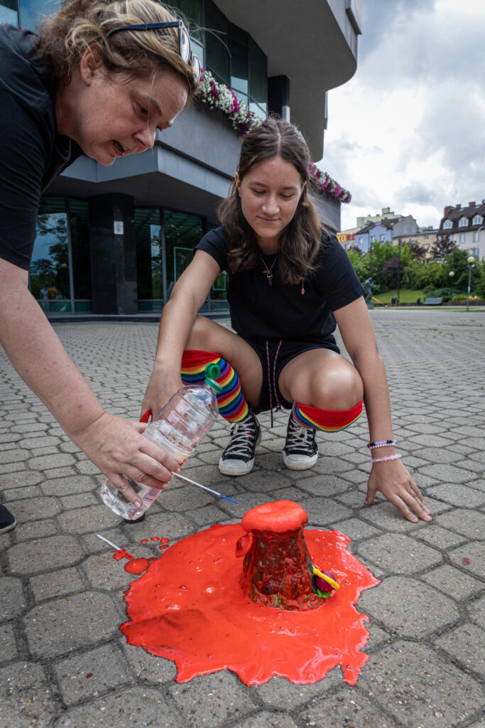 Zdjęcie. Dziewczyna kuca nad swoim wulkanem Nad nią stoi kobieta z plastikową butelką w ręce. Wlewa coś do krateru. Z wulkanu wylewa się intensywnie czerwona piana i rozlewa się na chodnik przed KCK.