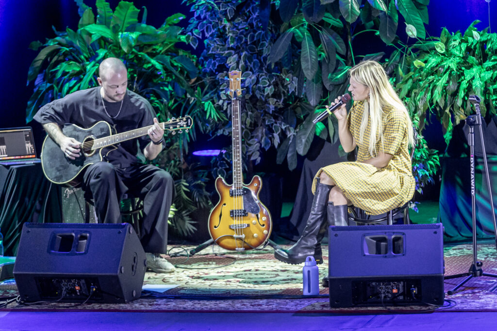 Zdjęcie. Dwójka ludzi - kobieta i mężczyzna siedzą na scenie na krzesłach. Blondynka z mikrofonem patrzy w kierunku gitarzysty. Za nimi duże zielone kwiaty.