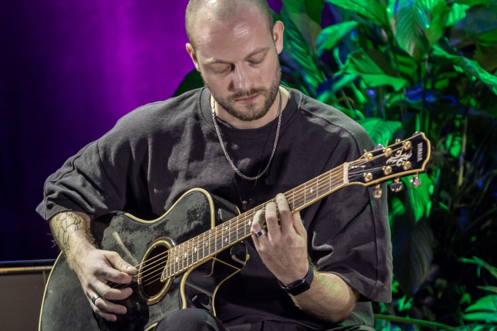 Zdjęcie. Mężczyzna gra na gitarze. To łysiejący mężczyzna z ciemna brodą i wąsami w czarnej podkoszulce.