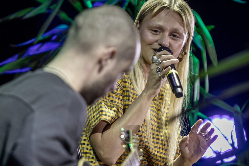 Zdjęcie. Kobieta śpiewa do mikrofonu. Ma długie rozpuszczone blond włosy, sukienkę żółtą w drobne niebieską kratkę. Siedzi na krześle. To Rosalie. Patrzy w kierunku gitarzysty.